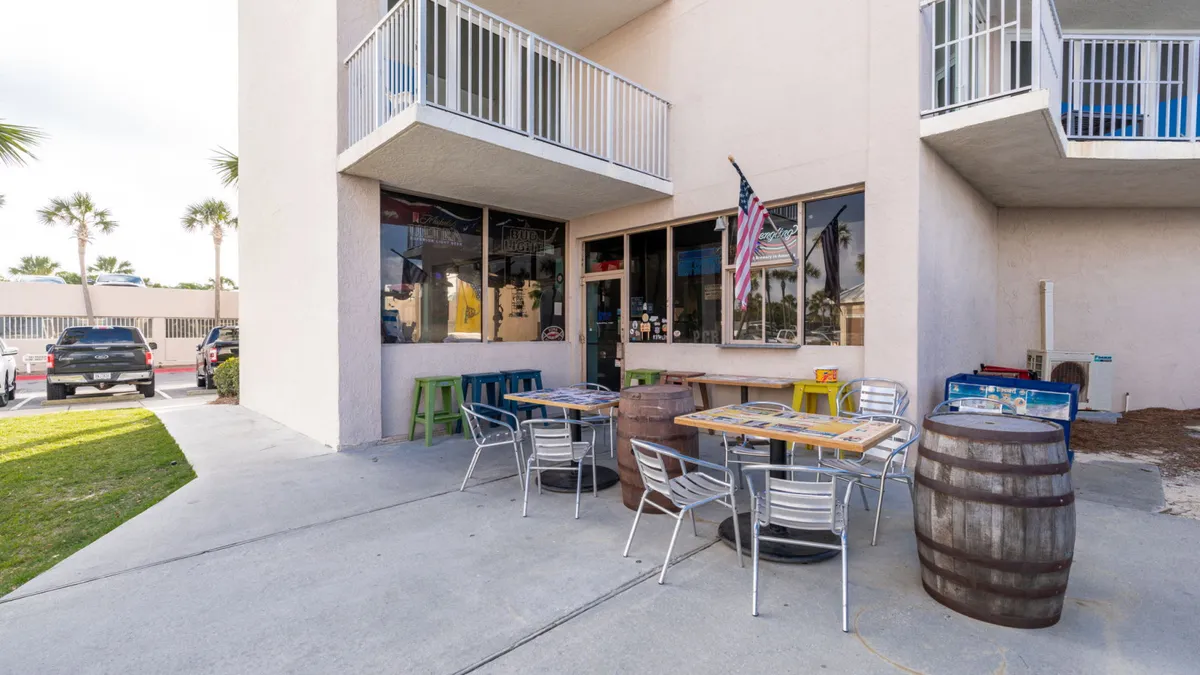 A lively outdoor seating area with colorful tables and barrels used as tables, perfect for casual dining and socializing. The tiki bar is visible, exuding a festive vibe ideal for enjoying bites and drinks after a swim or game.