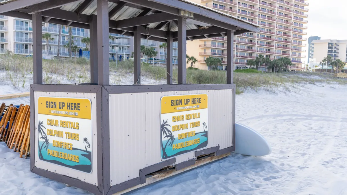 An inviting beachfront scene featuring a beach hut, fun signage, surfboard, and sandy beach extending toward distant condo buildings. The mood is vibrant and outdoorsy, capturing opportunities for beach activities, wildlife encounters, and enjoying gorgeous sunset views.