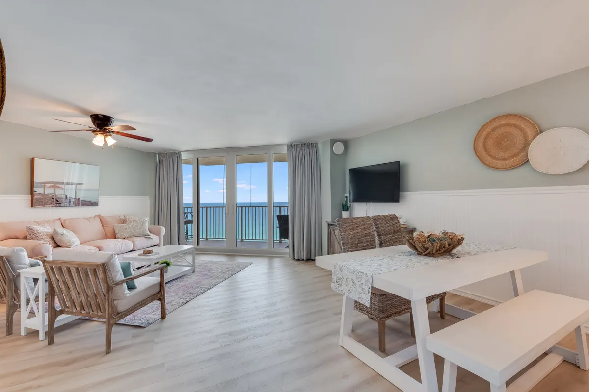 A bright, open-plan living and dining area featuring a sofa with pastel cushions, a dining table with woven chairs, and large windows offering unobstructed ocean views. The space is airy and designed for both relaxation and social gatherings, reinforcing the coastal ambience.
