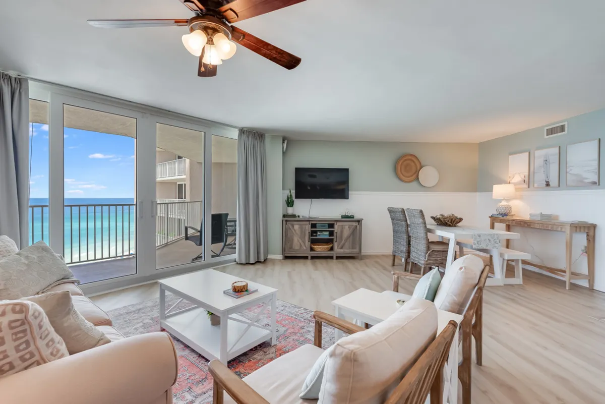 The image showcases a bright, open-plan living and dining area with large sliding glass doors framing sweeping Gulf-front ocean views. The comfortable seating area features a neutral-toned sofa and armchairs, while a dining table with wicker chairs invites gathering. The space flows seamlessly onto a private balcony, perfect for soaking in sunsets over the water.