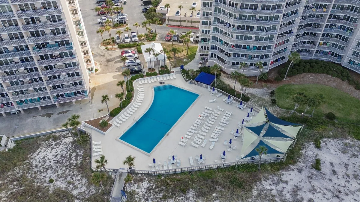 A large rectangular community pool is bordered by rows of lounge chairs and shaded by triangular canopy structures. High-rise condo buildings overlook the pool, while a sandy area and beach access path beckon in the background, tying together poolside relaxation and direct access to the Gulf.