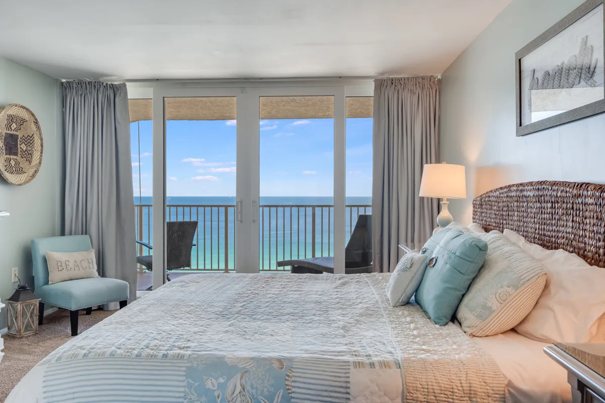 This image presents the primary king suite, featuring a large bed with a woven rattan headboard and pastel bedding. Floor-to-ceiling glass doors open onto a private balcony, providing an expansive view of the Gulf of Mexico. A comfortable accent chair with a beach-themed pillow completes the serene, inviting atmosphere.