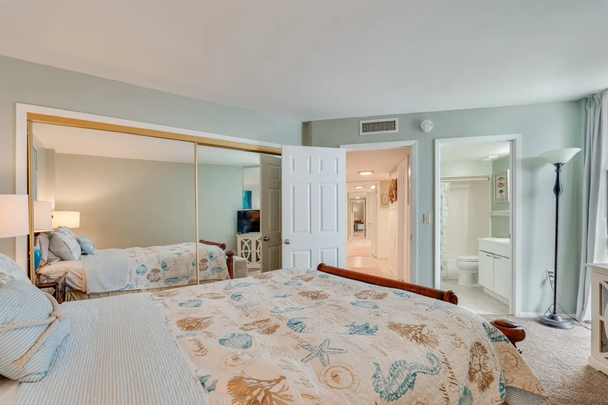 A cozy secondary bedroom is shown here, complete with a bed adorned in seashell and starfish patterned bedding, mirrored closet doors, and soft lighting from a floor lamp near the window. Open doorways reveal the adjoining bathroom and hallway, underscoring both comfort and convenient access to amenities.