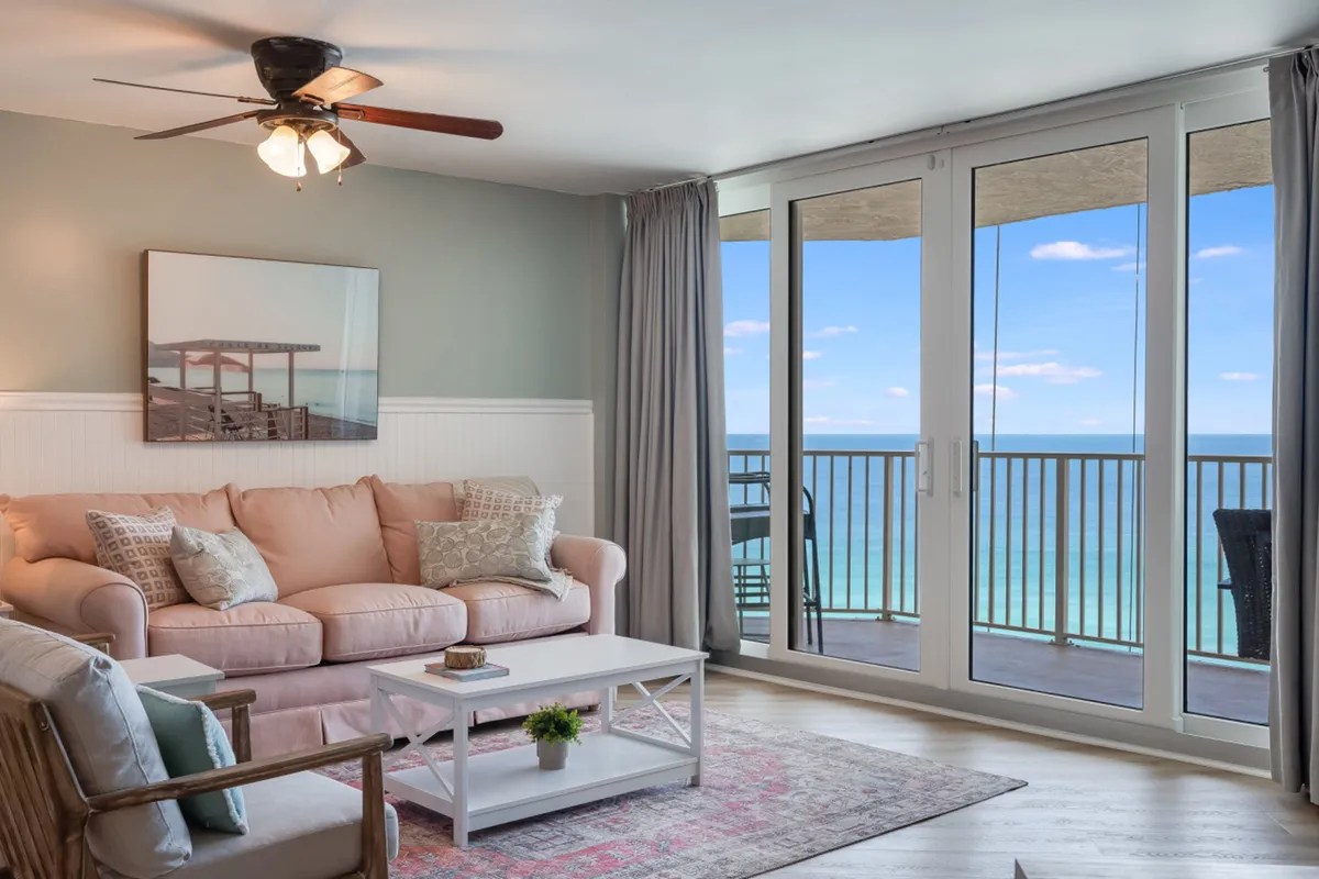 This image captures the living area, where a peach-colored sofa with patterned cushions faces large glass sliding doors framing a breathtaking Gulf view. A white coffee table and ceiling fan add to the inviting, home-like ambiance, with direct access to the balcony for ocean breezes.