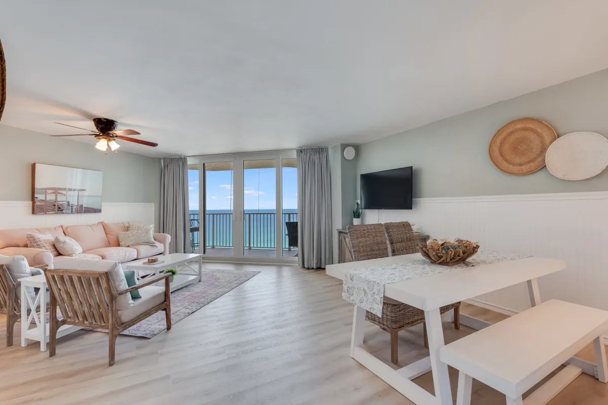 This interior photo features a comfortable peach sofa with accent pillows, a white dining table with bench and wicker chairs, and a wall-mounted TV with decorative baskets. Large glass doors lead out to a balcony offering beautiful Gulf views, uniting the living and dining zones with the outdoor space.