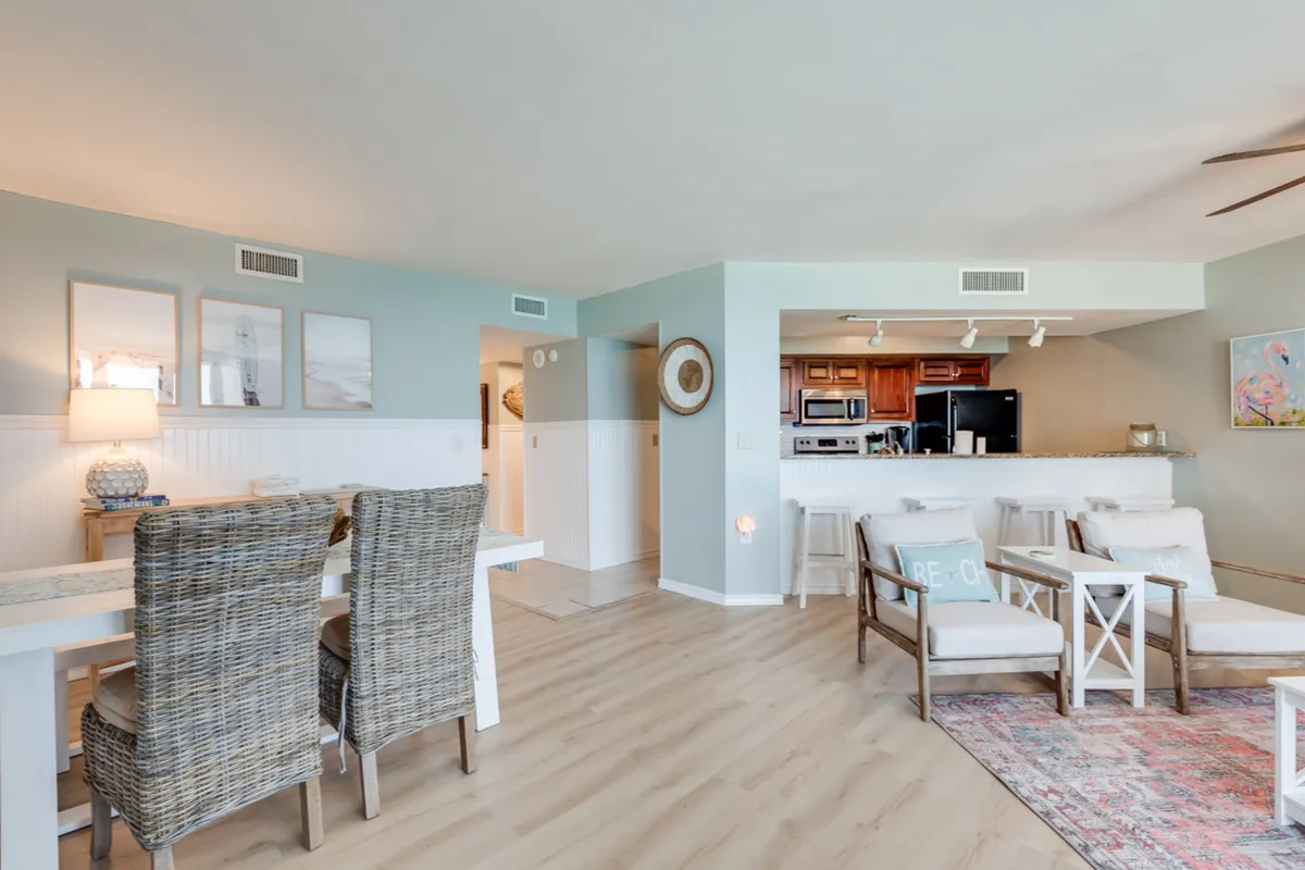 This image reveals an inviting open-concept space encompassing the dining area with wicker chairs, a living room with a 'Beach' pillow, and a modern kitchen outfitted with stainless steel appliances. The soft blue and white color scheme and coastal decor tie the kitchen and dining area together, reinforcing the relaxed vacation vibe. The kitchen is visible in the background, highlighting its modern features.