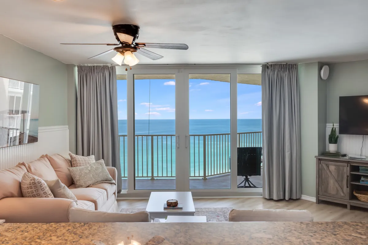 This image captures a spacious living room with a comfortable beige sectional sofa, patterned pillows, a mounted flat screen TV on a rustic wooden console, and large glass sliding doors that open to a balcony with sweeping Gulf views. Natural light fills the space, highlighting the seamless connection between the cozy living area and the outdoor balcony.
