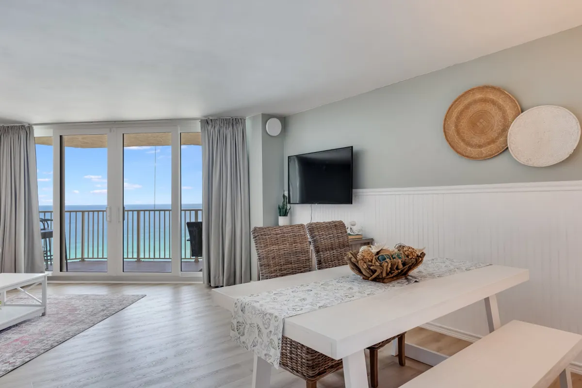 The photo showcases a bright dining area with a white table, woven chairs, and a decorative centerpiece. Large glass doors open to a balcony with Gulf views, and a wall-mounted flat screen TV and woven baskets add a coastal touch to the room. The scene is filled with natural light and provides an inviting space for meals and socializing.