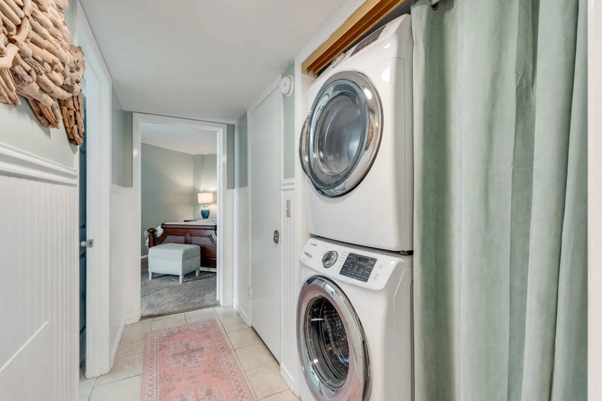 A stacked washer and dryer are nestled in a calm hallway adorned with decorative wall art and a soft area rug, leading toward a bedroom. The space feels welcoming and practical, emphasizing convenience for longer stays.