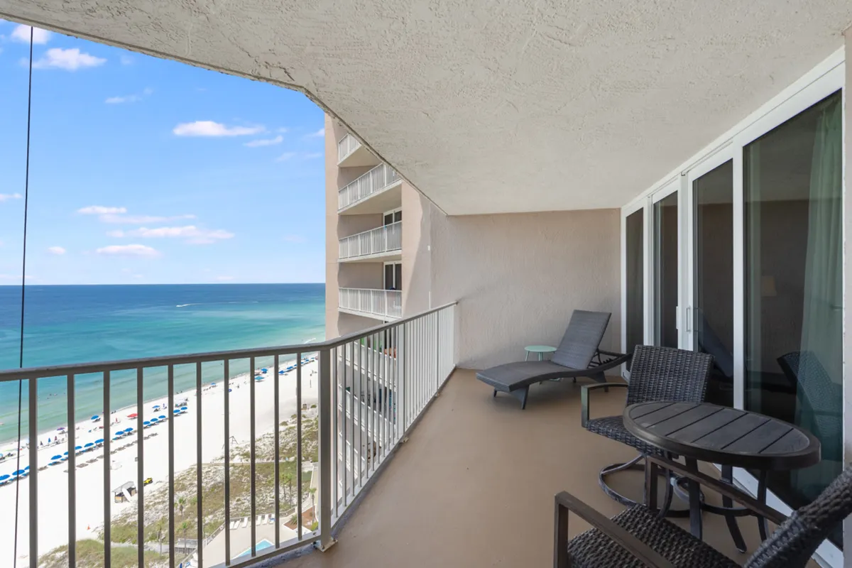 A private balcony overlooking the Gulf of Mexico, featuring an outdoor seating area with table and chairs and a lounger chair. Below, the white sand beach and blue umbrellas stretch along the shoreline.