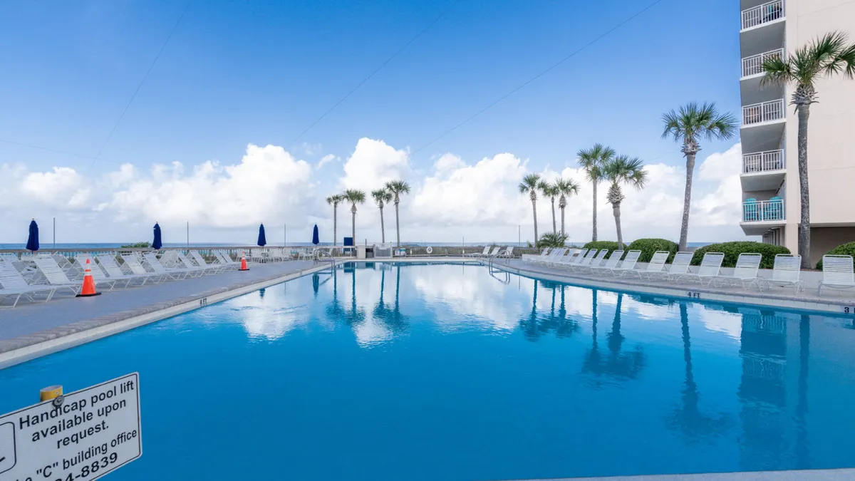 A large community swimming pool surrounded by palm trees and lounge chairs, with the ocean visible in the background. Blue umbrellas are set up poolside, and the entire scene is bathed in natural light, evoking a sense of leisure and relaxation.
