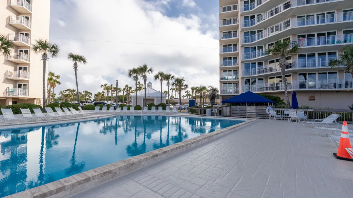 A large community swimming pool ringed by lounge chairs and tall palm trees, with a multi-story condo building and glimpses of the Gulf beach in the background. Several seating areas encourage relaxation and socializing.