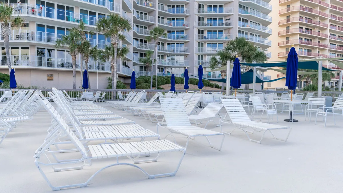 This image captures rows of white lounge chairs with blue closed umbrellas set around a tranquil pool, with tall palm trees and a modern condo building with balconies in the background. The scene is bright, inviting, and suggests a calm, leisurely vacation environment.