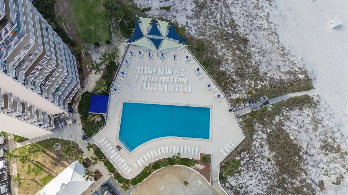 A large, sparkling community swimming pool surrounded by neat rows of white lounge chairs and shaded seating areas with blue and beige sails. The pool is adjacent to sandy beach dunes and lush vegetation, creating a seamless transition from poolside relaxation to beach exploration.