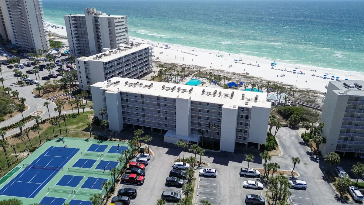 A vibrant exterior scene showcasing blue tennis and pickleball courts in the foreground, with a white multi-story condo building behind. The sandy beach and turquoise Gulf waters are visible beyond, and a parking area with vehicles sits to the side.