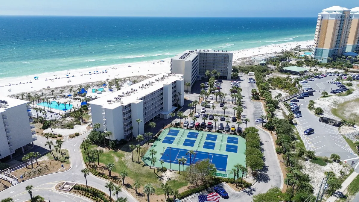 A dynamic aerial view showing the beachfront with turquoise Gulf waters, community pools surrounded by sun loungers, pickleball and tennis courts, and modern condo buildings. The image captures the variety of resort amenities and the vibrant, active atmosphere of the community.