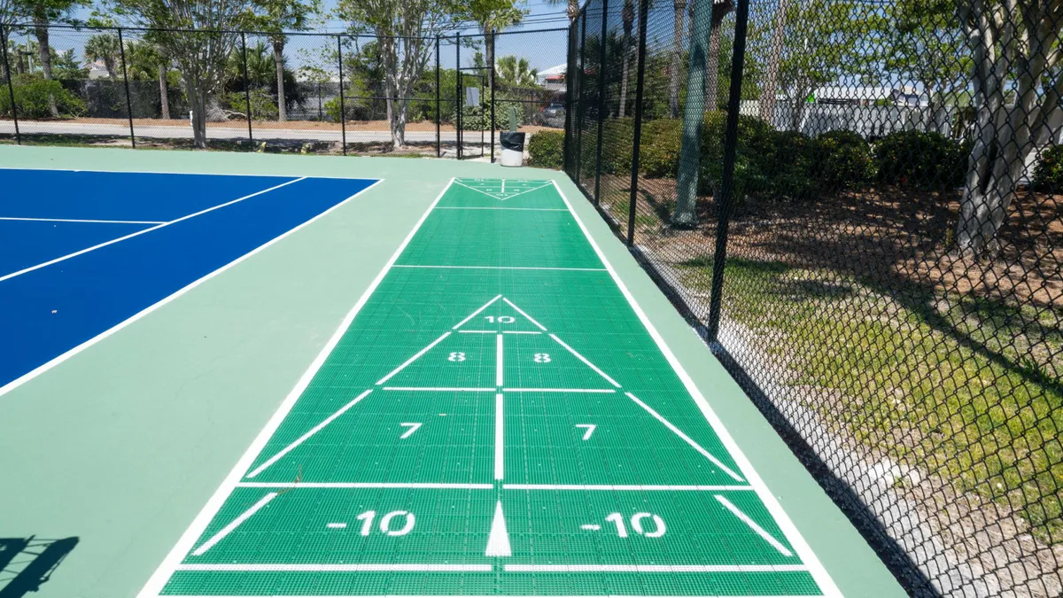 A lively shuffleboard court with clearly marked zones sits alongside blue-colored tennis or pickleball courts, surrounded by lush greenery and trees. The outdoor setting invites friendly competition and family togetherness in the fresh coastal air.