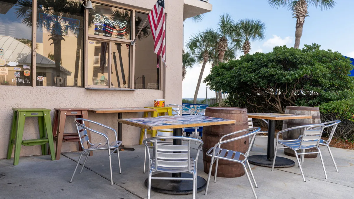 Outdoor dining area with metal chairs and wooden barrel tables, accented by an American flag and lush palm trees. The setup suggests a lively, sociable atmosphere ideal for enjoying drinks or meals from the tiki bar.