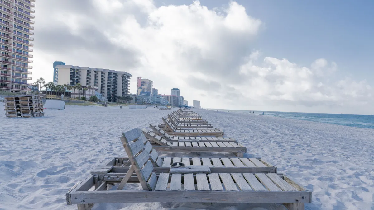 A row of wooden lounge chairs lines a pristine, white sandy beach with the Gulf of Mexico stretching to the horizon. High-rise condos and buildings border the beachfront, and the blue sky is dotted with clouds. The scene is peaceful and spacious, ideal for long beach walks or quiet moments.