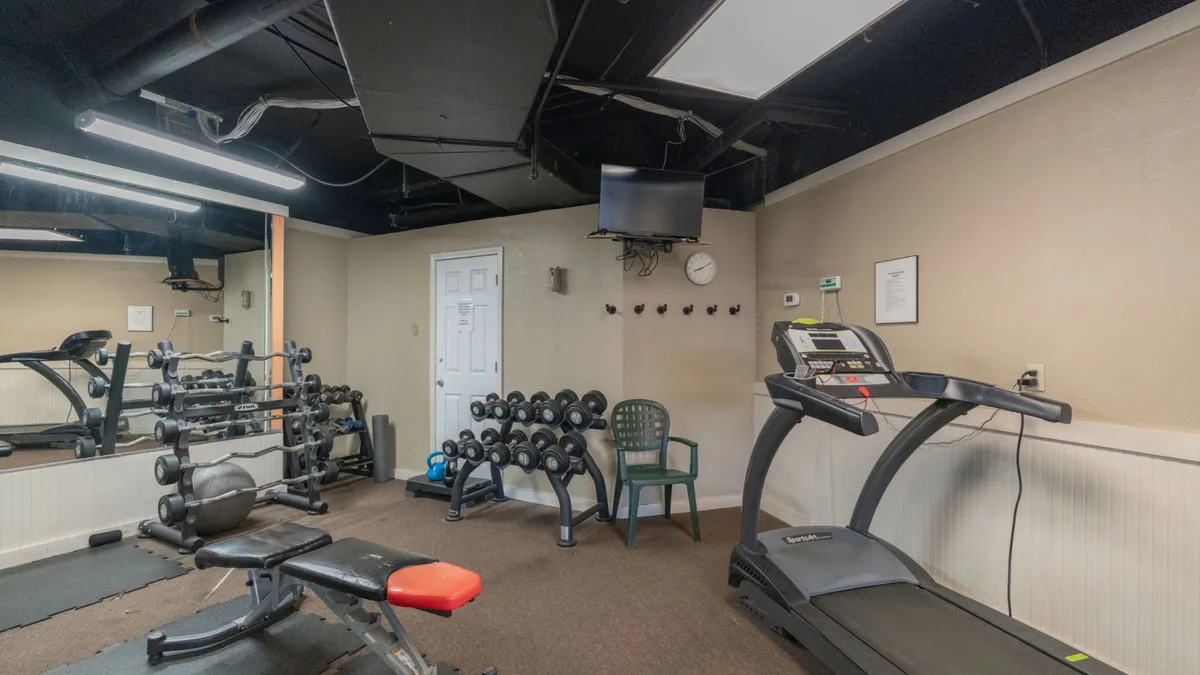 A bright, well-equipped fitness center featuring a treadmill, weight bench with red padding, dumbbell racks, and a wall-mounted television. The environment is motivating and suitable for both cardio and strength workouts.