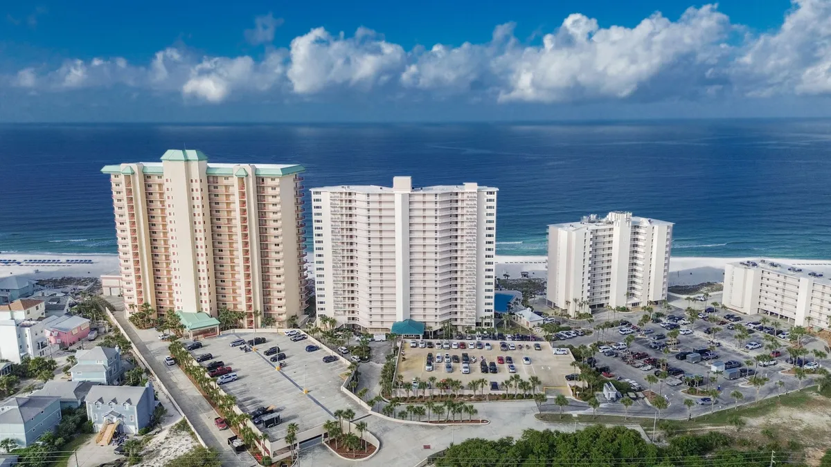 Three high-rise condominium buildings stand prominently near the shoreline, framed by a wide sandy Gulf beach and turquoise waters. The image also reveals well-maintained parking lots and landscaped resort surroundings, conveying both the proximity to the beach and the scale of the resort complex.