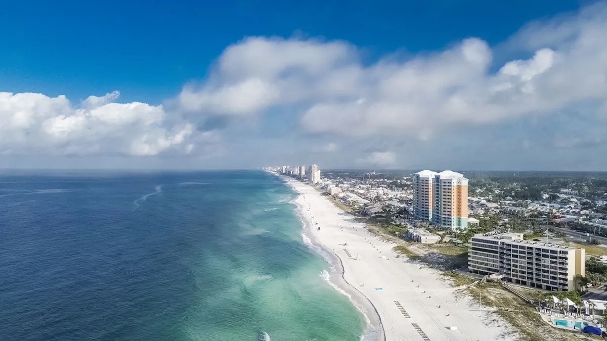 A sweeping view of a long stretch of white sandy beach meeting the clear blue-green waters of the Gulf, with high-rise beachfront condos and a wide open sky overhead. The scene is calm and inviting, with people enjoying the beach, swimming, and sunbathing, capturing the essence of a family-friendly coastal escape.